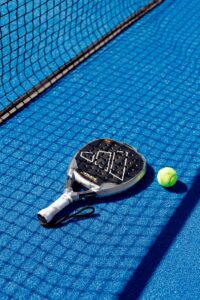 Padel racket and ball resting on a vibrant blue court, next to a net, under bright daylight.